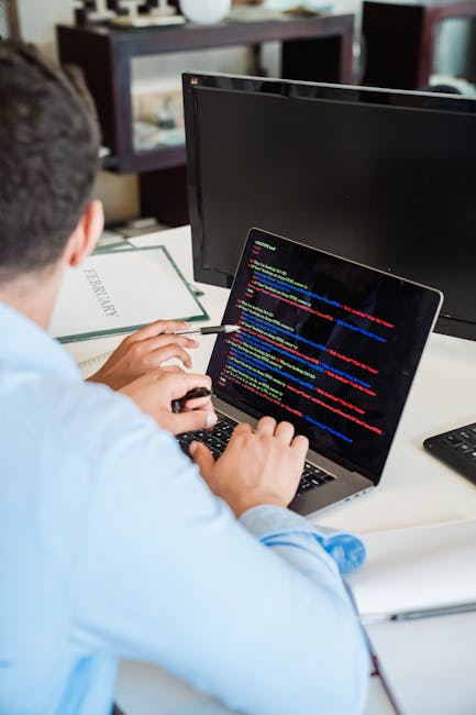 A person working at a desk with a laptop displaying lines of colourful code, situated in an indoor office environment. The individual is focused on the screen, holding a pen in one hand, with a book l
