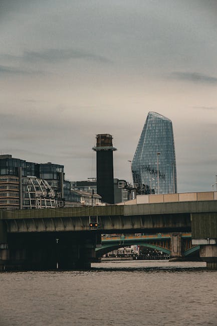 The image shows a cityscape viewed from across the River Thames, with an emphasis on urban landscaping elements. In the foreground, a concrete bridge spans the river, featuring a textured, slightly we