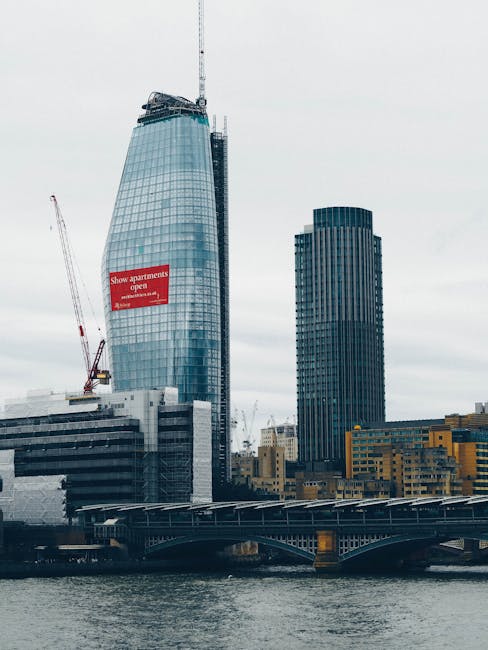 The image shows a cityscape viewed from across the River Thames, with an emphasis on urban landscaping elements. In the foreground, a concrete bridge spans the river, featuring a textured, slightly we