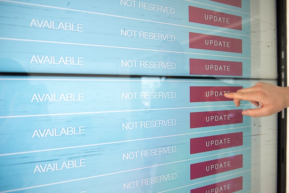 A person's hand is pointing at a digital scheduling display with multiple rows, each showing the status 'AVAILABLE' and 'NOT RESERVED' alongside red 'UPDATE' buttons. The background of the screen is l